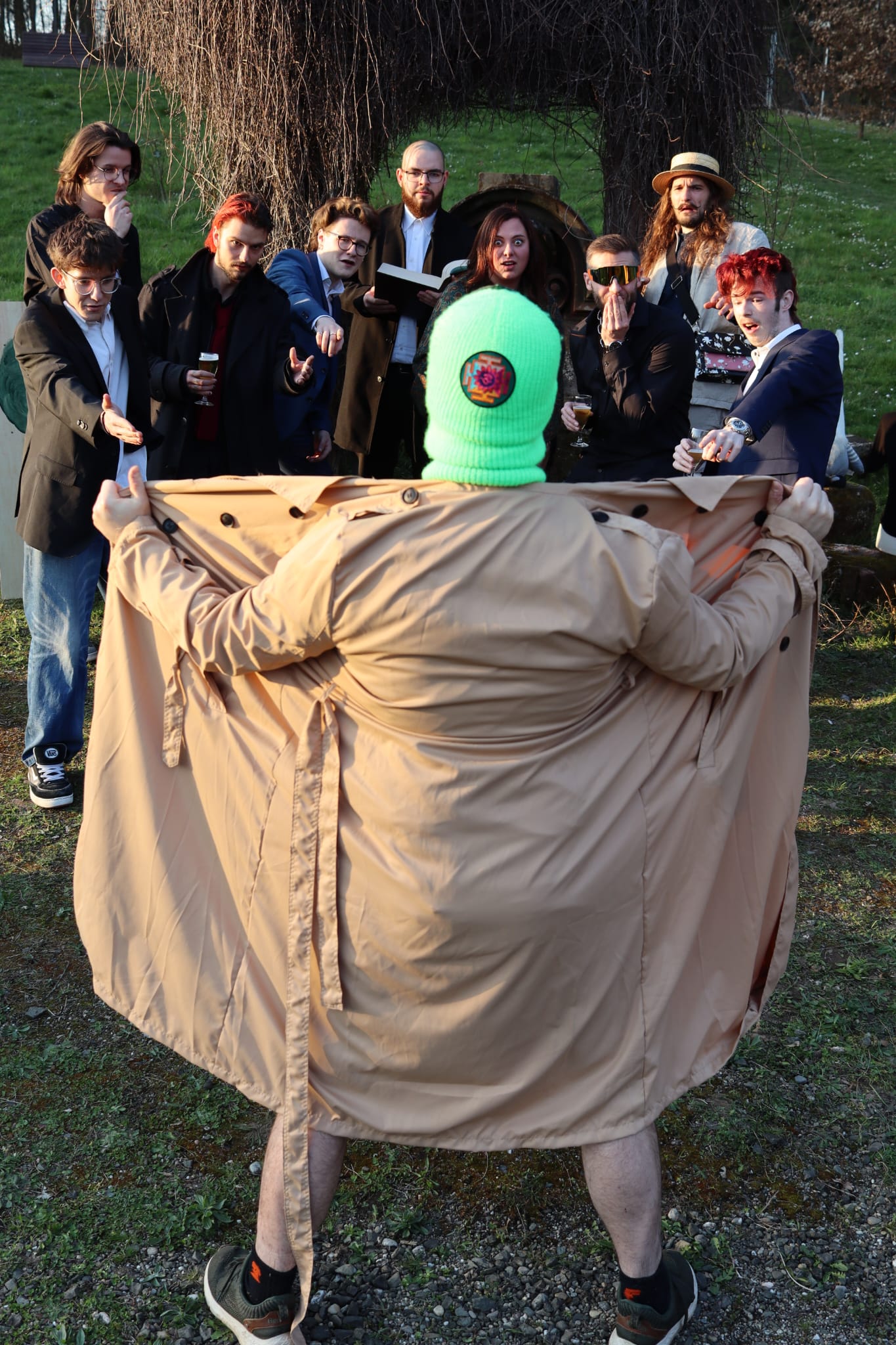 Der Mann mit der neongrünen Sturmhaube öffnet seinen Trenchcoat vor entsetztem Publikum
